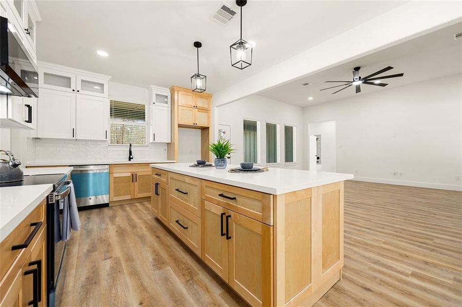 This photo showcases a modern, open-concept kitchen and living area. The kitchen features a large island with light wood cabinetry, white countertops, and pendant lighting. Hardwood floors create a warm ambiance, and the space is bright with ample natural light.