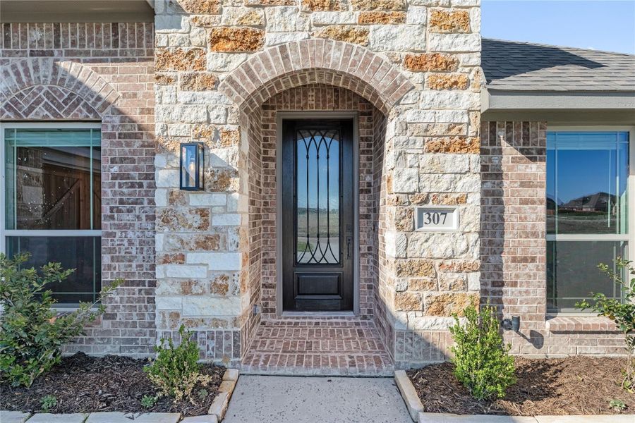 Home entrance with brick and stone arch, dark iron front door, and wall sconce light fixture Home entrance with brick and stone arch, dark iron front door, and wall sconce light fixture