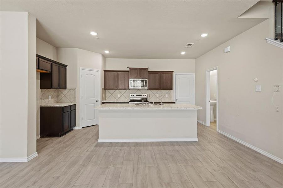 Kitchen with backsplash, light wood-style floors, a kitchen island with sink, dark brown cabinets, and light stone counters Kitchen with backsplash, light wood-style floors, a kitchen island with sink, dark brown cabinets, and light stone counters