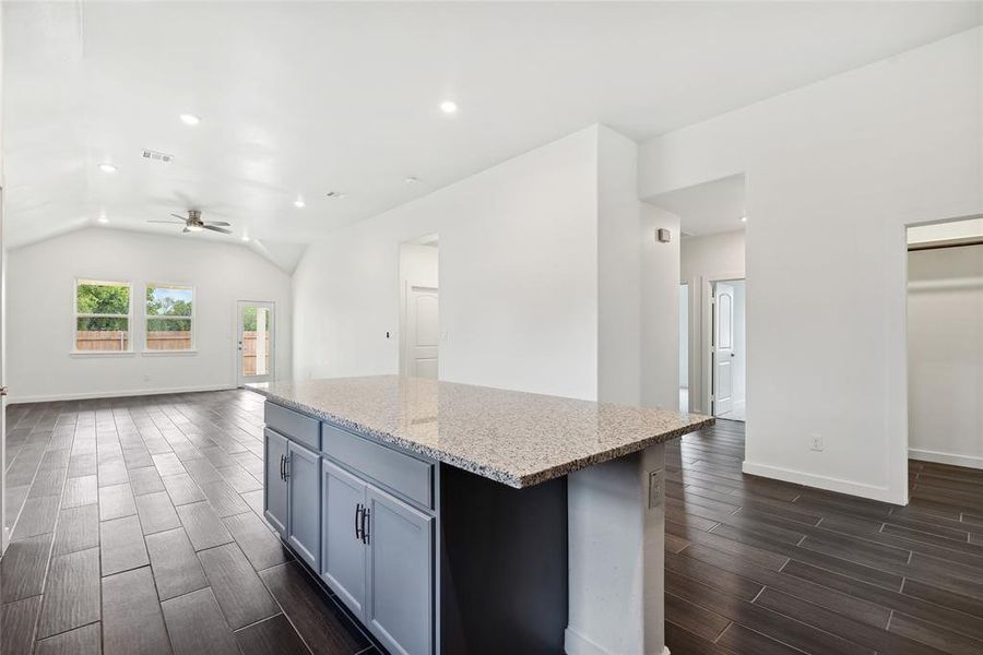 Kitchen with vaulted ceiling, light stone counters, dark wood-type flooring, ceiling fan, and a kitchen island