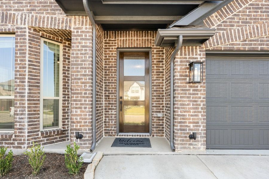 Brick home entrance with wood and glass front door, welcome mat, and gray garage door nearby. Brick home entrance with wood and glass front door, welcome mat, and gray garage door nearby.