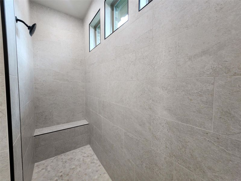 Walk-in shower featuring light-toned tile walls, a built-in bench, pebbled flooring, a black shower head, and transom windows