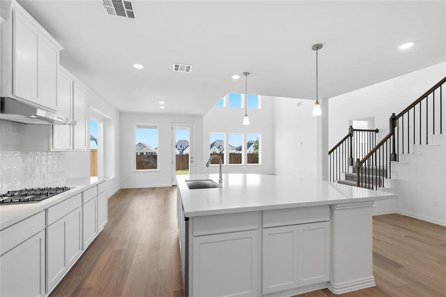 Kitchen featuring white cabinets, light wood-type flooring, an island with sink, decorative light fixtures, and stainless steel gas cooktop