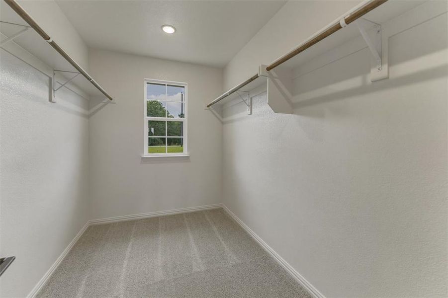 Walk-in closet featuring light gray carpeting, two clothing rods with shelving, a single recessed ceiling light, and a window with white trim Walk-in closet featuring light gray carpeting, two clothing rods with shelving, a single recessed ceiling light, and a window with white trim