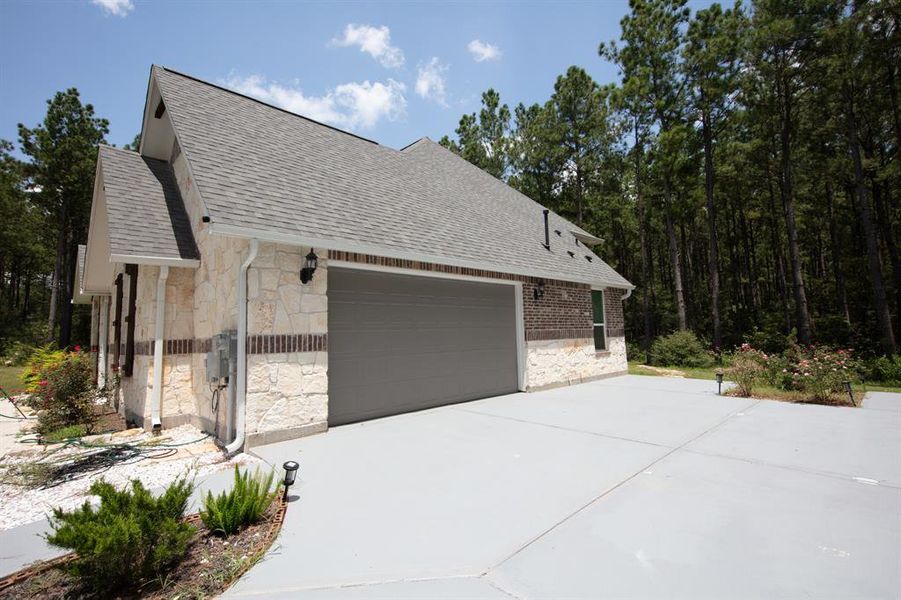 This photo shows a modern home with a stone and brick exterior, featuring a spacious two-car garage. It is set against a backdrop of tall trees, providing a natural and private setting. The driveway is large, and the landscaping is simple, with some greenery near the entrance.