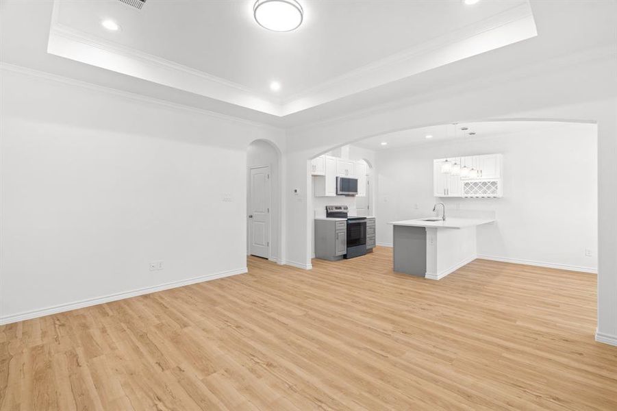 Unfurnished living room featuring light wood-style floors, a raised ceiling, arched walkways, crown molding, and recessed lighting Unfurnished living room featuring light wood-style floors, a raised ceiling, arched walkways, crown molding, and recessed lighting