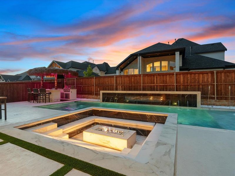 Backyard oasis featuring a sunken fire pit lounge with built-in seating, a swimming pool with integrated water features, and an outdoor kitchen area with a thatched-roof bar Backyard oasis featuring a sunken fire pit lounge with built-in seating, a swimming pool with integrated water features, and an outdoor kitchen area with a thatched-roof bar