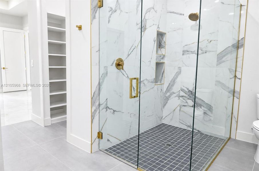 Walk-in shower featuring gold-toned hardware, floor-to-ceiling tiled walls with marbled pattern, recessed wall shelving, and a glass enclosure, complemented by built-in wall cabinetry