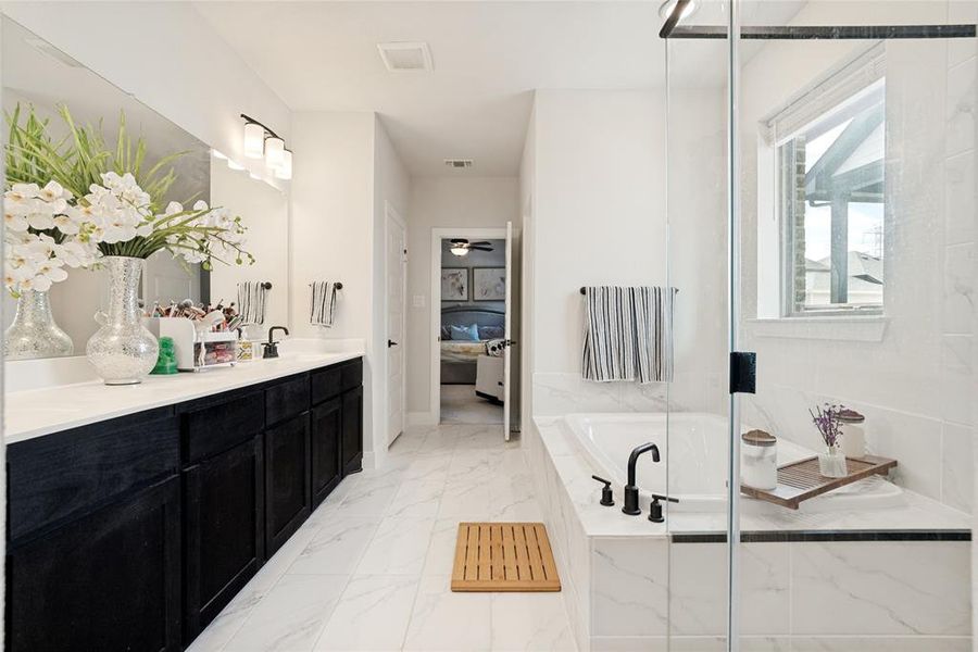 Bathroom featuring a dual vanity with black cabinetry and white countertops, a built-in tub with a matte black faucet, a glass-enclosed shower, and white tile flooring