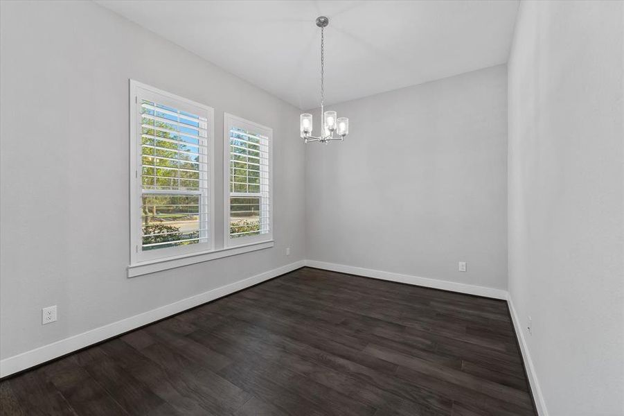 This is a bright, dining room/ or home office with neutral walls, laminate wood flooring, and a modern hanging light fixture. There are two large windows with plantation shutters, offering ample natural light and a view of greenery outside.