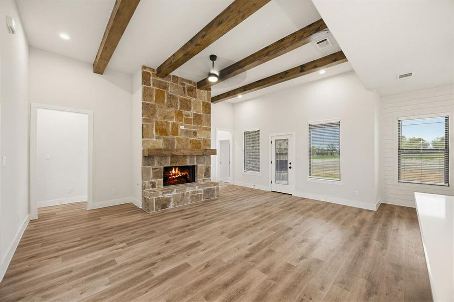 Vaulted ceilings featuring exposed wood beams, a floor-to-ceiling stone fireplace with a wood mantle, wood-finish flooring, and multiple windows providing natural light