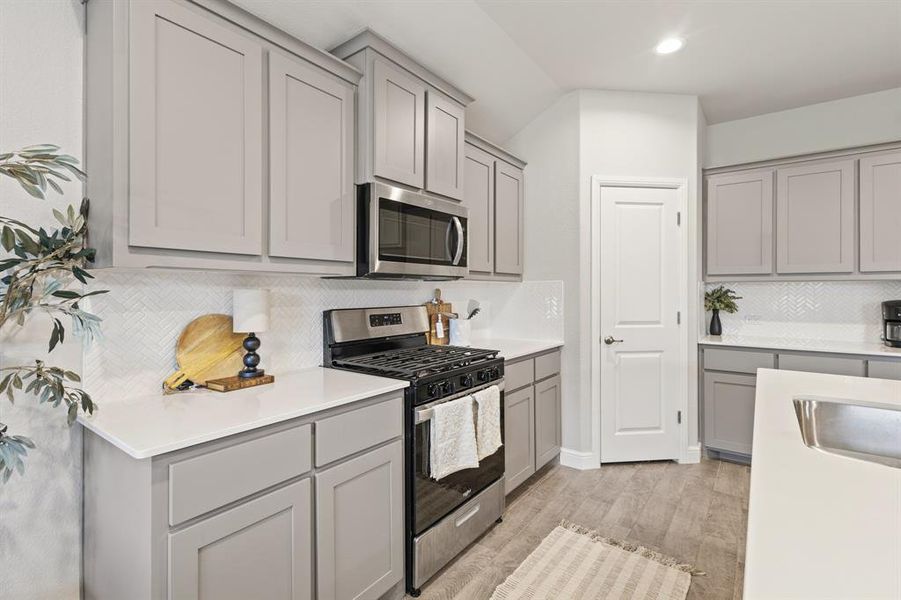Contemporary kitchen featuring light gray cabinetry, stainless steel appliances, white countertops, a white herringbone tile backsplash, and wood-finish flooring Contemporary kitchen featuring light gray cabinetry, stainless steel appliances, white countertops, a white herringbone tile backsplash, and wood-finish flooring