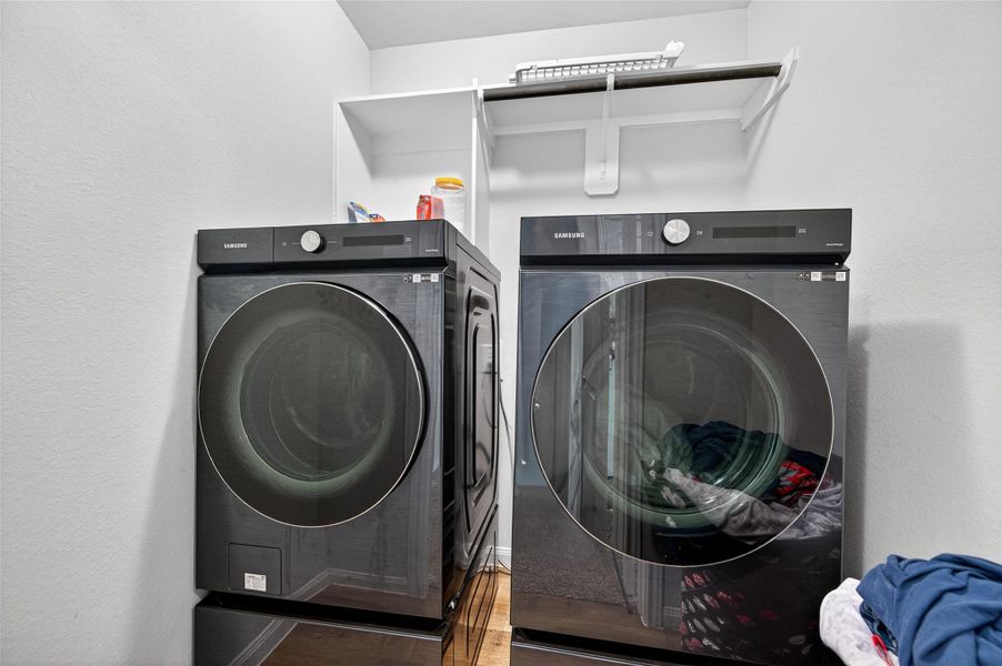 This photo shows a laundry room featuring modern Samsung washer and dryer units with a clean, minimalist setup. There's a shelf above for storage, set against a neutral wall, providing a functional and organized space.