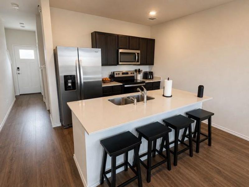 Open-concept kitchen featuring an island with an integrated sink and bar seating, stainless steel appliances, dark wood-finish cabinetry, and wood-finish flooring throughout Open-concept kitchen featuring an island with an integrated sink and bar seating, stainless steel appliances, dark wood-finish cabinetry, and wood-finish flooring throughout