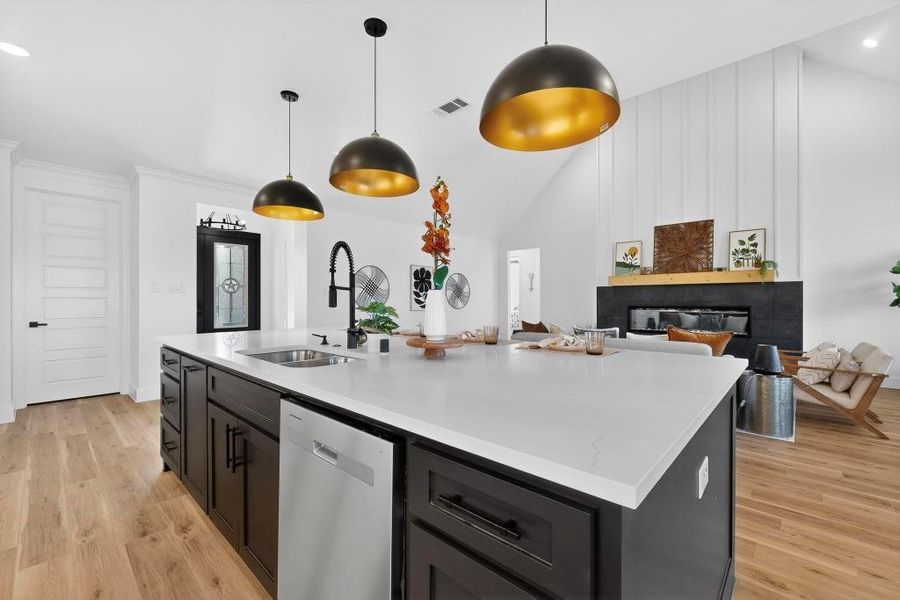 Kitchen featuring light wood finished floors, dishwasher, a glass covered fireplace, decorative light fixtures, and a kitchen island with sink Kitchen featuring light wood finished floors, dishwasher, a glass covered fireplace, decorative light fixtures, and a kitchen island with sink