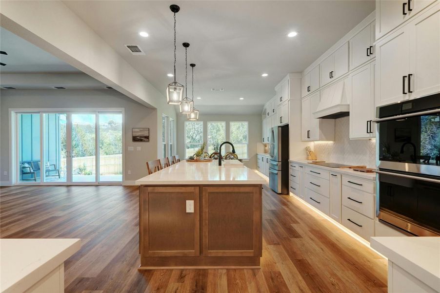 Modern kitchen featuring an expansive center island with seating, pendant lighting, white shaker-style cabinetry, stainless steel appliances, and wood-finish flooring