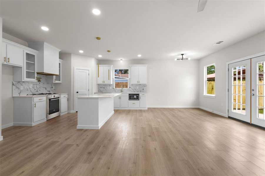 Open-concept living space featuring wood-finish flooring throughout, a modern kitchen with white cabinetry and an island, a gas range with a contemporary range hood, and double French doors to the exterior