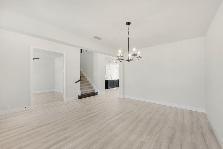 Open dining area with light wood floors, chandelier, staircase, and kitchen visible in background