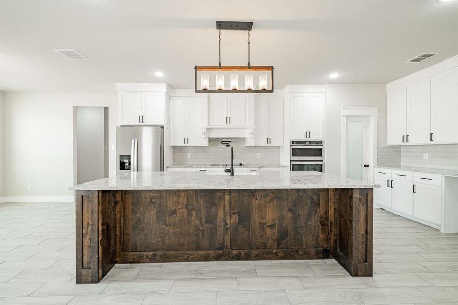 Kitchen featuring a large island, white cabinetry, hanging light fixtures, appliances with stainless steel finishes including refrigerator, wine glass rack and wine/soda frig (to be installed) and tasteful backsplash Kitchen featuring a large island, white cabinetry, hanging light fixtures, appliances with stainless steel finishes including refrigerator, wine glass rack and wine/soda frig (to be installed) and tasteful backsplash