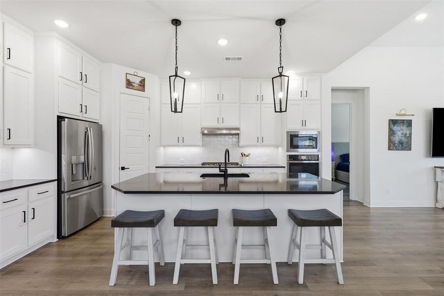 Open-concept kitchen featuring white cabinetry, a large central island with dark countertops, stainless steel appliances, and wood-finish flooring Open-concept kitchen featuring white cabinetry, a large central island with dark countertops, stainless steel appliances, and wood-finish flooring