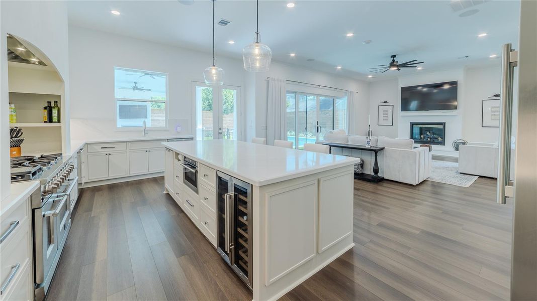 This modern open-concept kitchen and living area features sleek hardwood floors, a large central island with ample storage, stainless steel appliances, and elegant pendant lighting. The living space includes a cozy fireplace and large windows offering plenty of natural light and a view of the backyard.