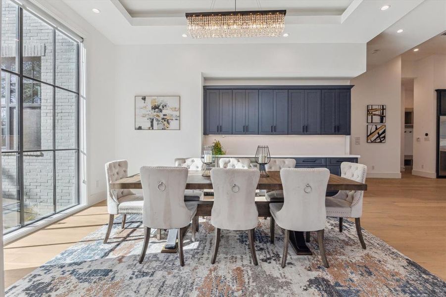 This formal dining room features a spacious layout with a large window offering natural light and views of the outdoors. The coffered ceiling adds depth and elegance, complemented by recessed lighting and a central chandelier for ambiance. Along the back wall, built-in navy cabinetry with sleek hardware provides ample storage and a polished backdrop. The light wood flooring ties the space together, creating a warm and inviting atmosphere.