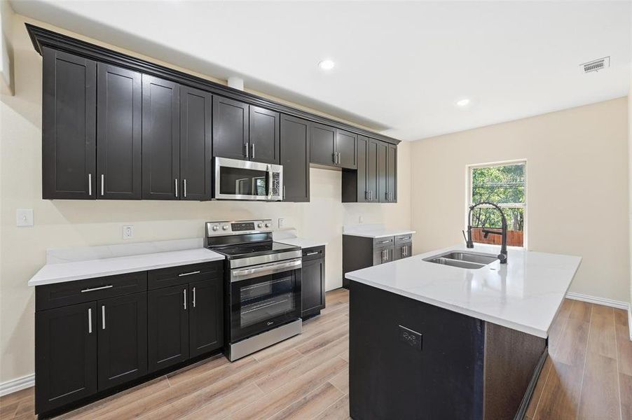Kitchen featuring stainless steel appliances, dark cabinetry, light stone counters, light wood finished floors, and a center island with sink Kitchen featuring stainless steel appliances, dark cabinetry, light stone counters, light wood finished floors, and a center island with sink
