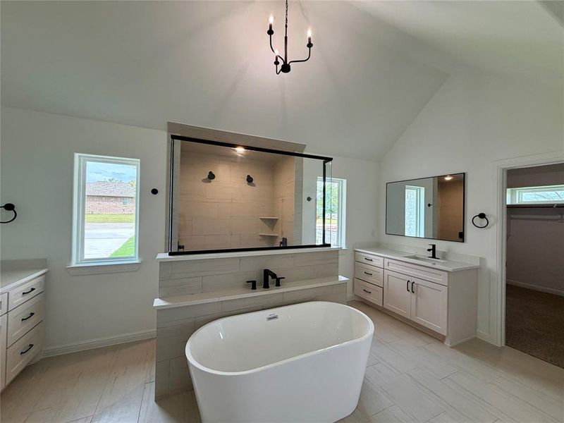 Spacious bathroom featuring a freestanding soaking tub, walk-in shower with glass enclosure, dual vanity areas with white cabinetry, a vaulted ceiling, and light-toned tile flooring