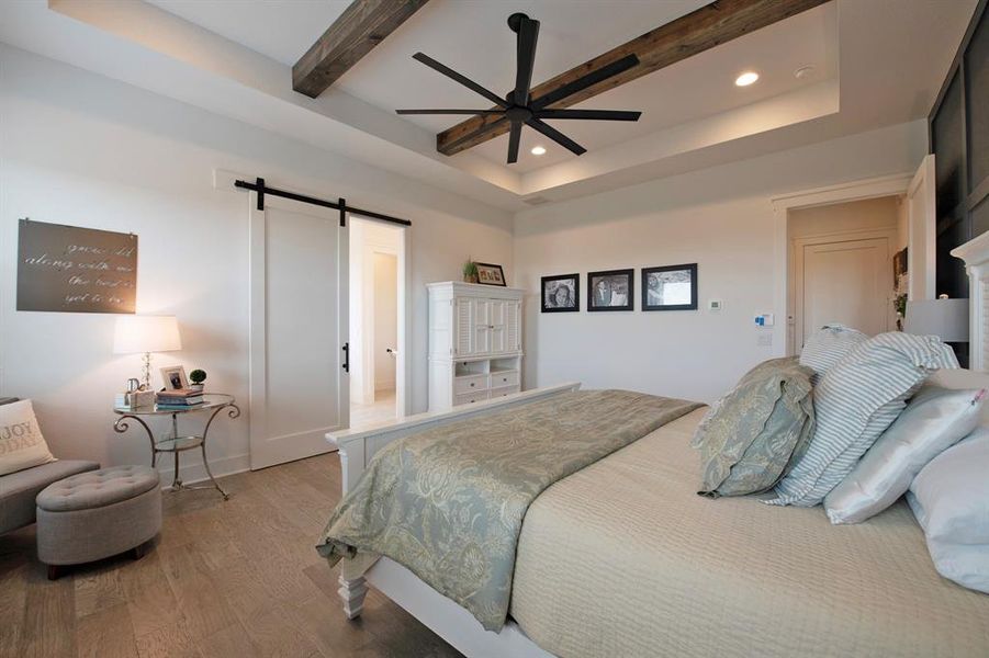 Bedroom with a barn door, wood finished floors, a raised ceiling, beam ceiling, and recessed lighting
