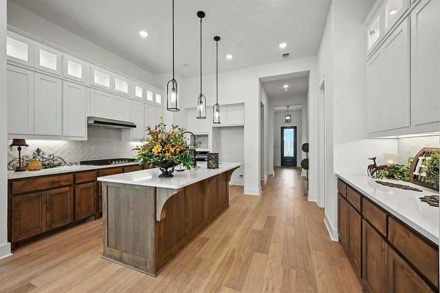 Kitchen with backsplash, glass insert cabinets, an island with sink, hanging light fixtures, and light wood-style floors