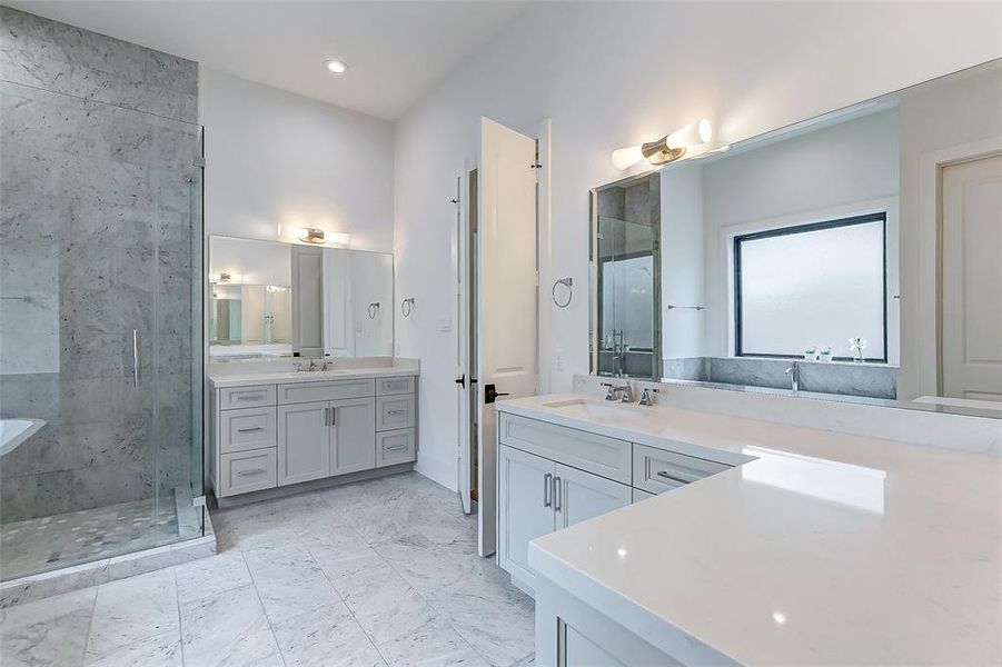 This bathroom features a spacious layout with elegant marble flooring and quartz countertops, dual sinks, and a large glass-enclosed shower. It includes ample storage with sleek cabinetry and a large mirror, providing a bright and modern feel.