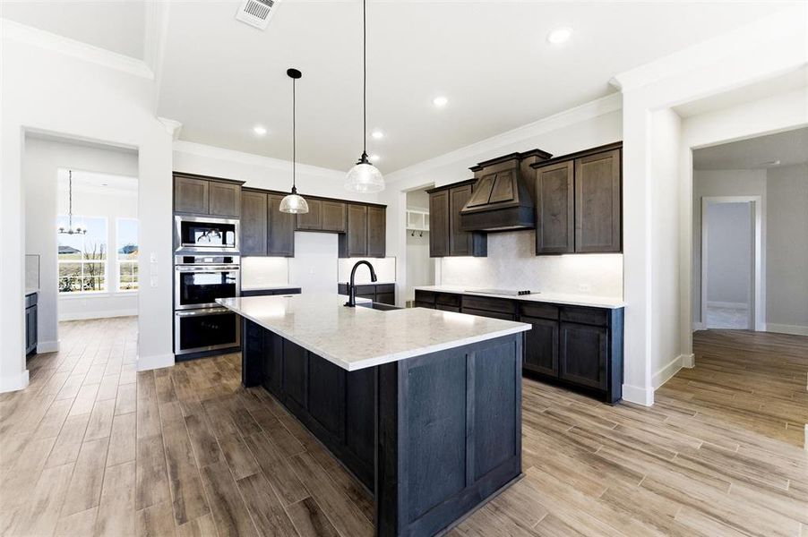 Kitchen with ornamental molding, decorative light fixtures, light stone countertops, a center island with sink, and light wood-style floors