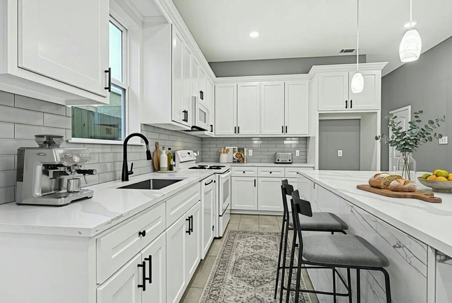 This kitchen features a modern design with white cabinetry and countertops, complemented by a gray subway tile backsplash. It includes appliances, a sleek black faucet, and a breakfast bar with seating. Pendant lights add a stylish touch, creating a bright and inviting space.