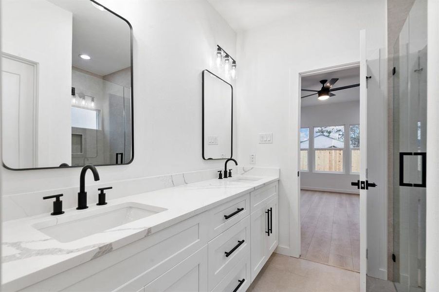 This photo showcases a modern bathroom with a double vanity featuring sleek black fixtures and large mirrors. The room is bright with white walls and a view into an adjacent room with a ceiling fan and large windows. This photo showcases a modern bathroom with a double vanity featuring sleek black fixtures and large mirrors. The room is bright with white walls and a view into an adjacent room with a ceiling fan and large windows.