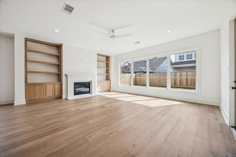 Natural light pours through oversized picture windows, casting warmth across wide plank hardwood floors and the clean lined fireplace. Custom wood built ins add texture and storage while framing views of the private backyard and covered veranda.