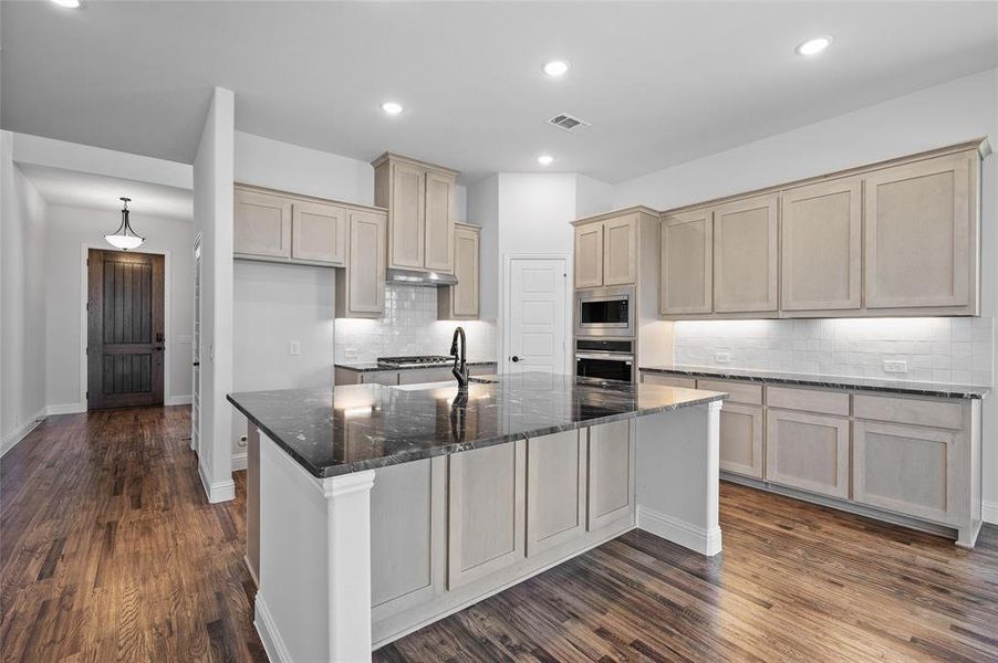 Kitchen featuring dark stone countertops, decorative backsplash, dark wood-type flooring, a kitchen island with sink, and appliances with stainless steel finishes