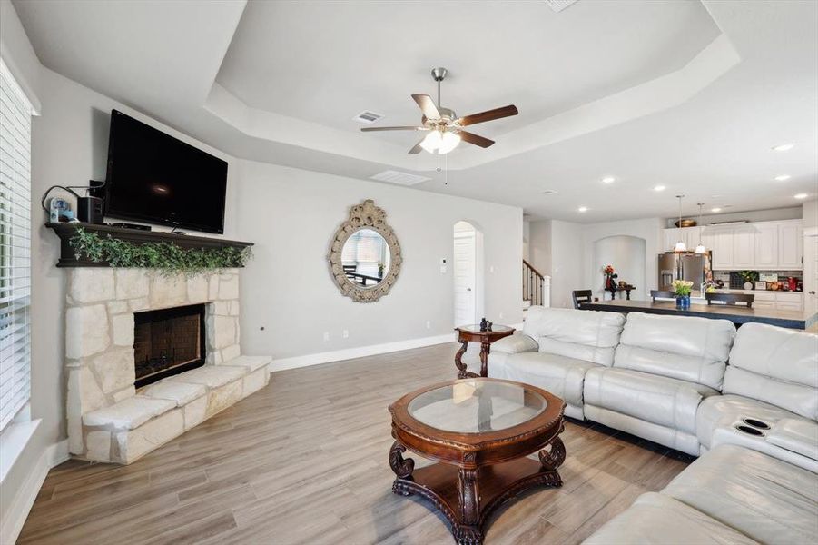 Living room with light wood-type flooring, ceiling fan, a stone fireplace, and a raised ceiling