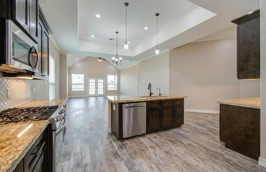 This is a spacious, modern kitchen with dark wood cabinetry, granite countertops, stainless steel appliances, and a central island. It opens to a large, well-lit living area with high ceilings and multiple windows, offering a bright and airy space. The flooring throughout is a wood-look tile.