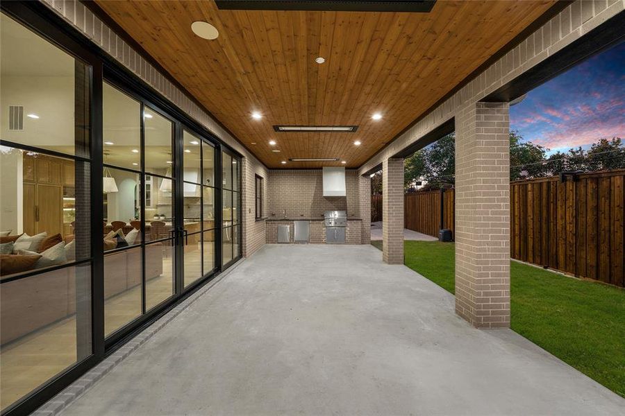Covered patio featuring an outdoor kitchen with built-in appliances, brick veneer accents, wood plank ceiling with recessed lighting, and expansive sliding glass doors