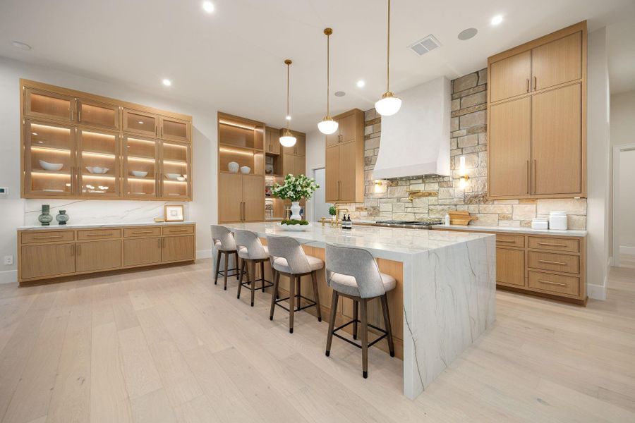 This beautifully designed kitchen showcases refined craftsmanship with custom cabinetry, integrated lighting, and a spacious island perfect for gathering. Sleek pendant lighting adds warmth and balance, while the expansive layout enhances flow and functionality. The standout feature is the illuminated glass-front cabinetry, offering both display and ambiance, creating a sophisticated yet inviting atmosphere ideal for hosting or daily living.