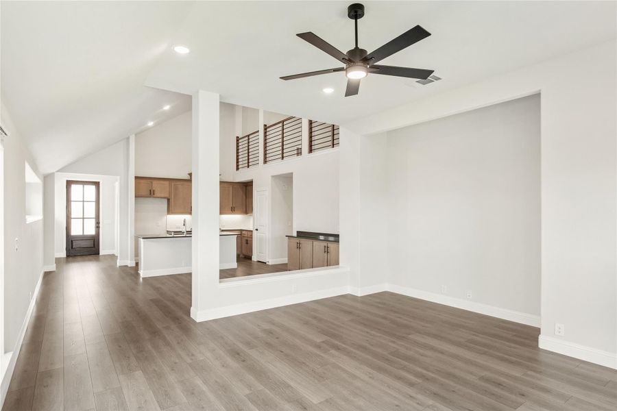 Open living area with vaulted ceiling, ceiling fan, hardwood floors, and open kitchen visible in background Open living area with vaulted ceiling, ceiling fan, hardwood floors, and open kitchen visible in background