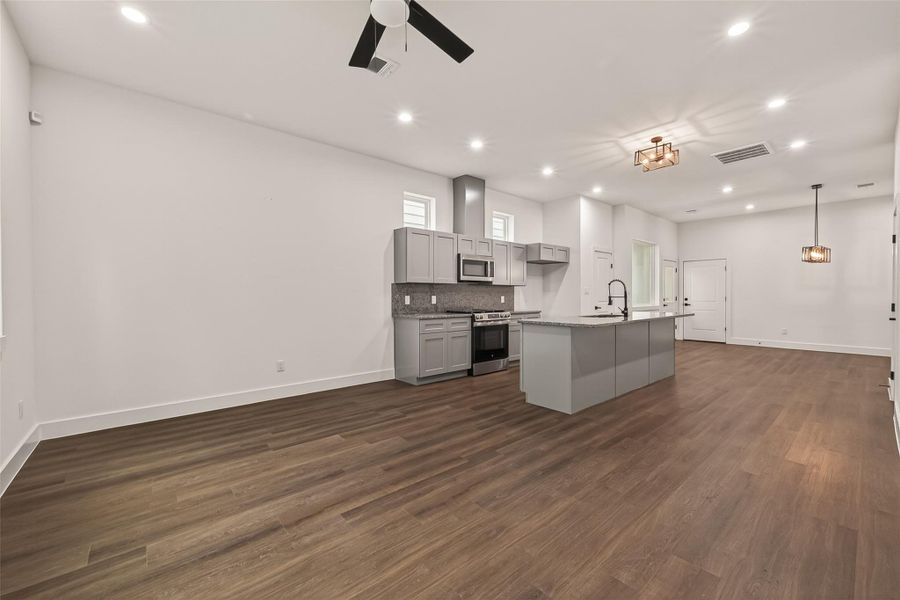 This photo showcases a spacious, open-concept living area with a modern kitchen featuring grey cabinets, stainless steel appliances, and a large island. The room has wood flooring, recessed lighting, and stylish ceiling fixtures, providing a clean and contemporary look. This photo showcases a spacious, open-concept living area with a modern kitchen featuring grey cabinets, stainless steel appliances, and a large island. The room has wood flooring, recessed lighting, and stylish ceiling fixtures, providing a clean and contemporary look.