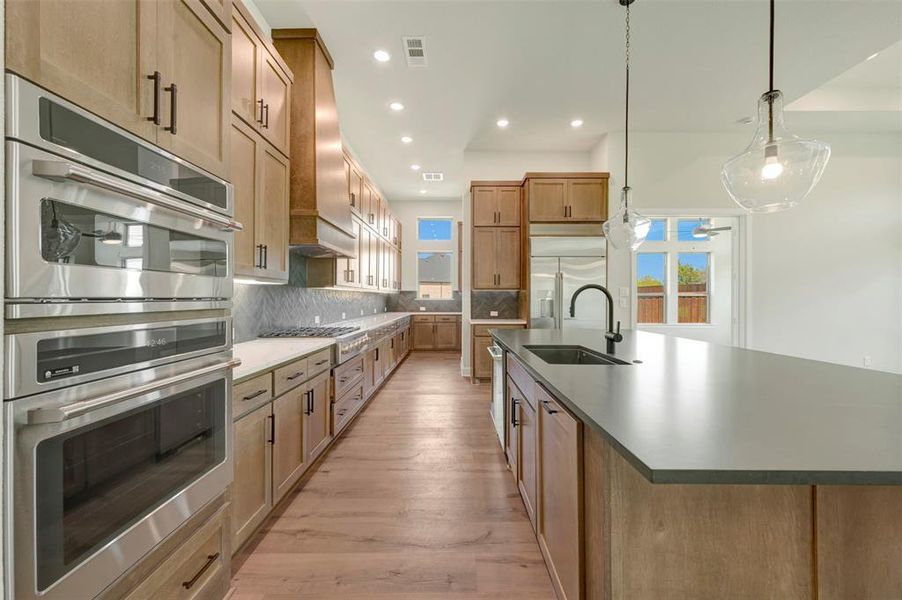 Kitchen featuring stainless steel appliances, a large island with sink, light wood-style floors, wood finish cabinets, and tasteful backsplash