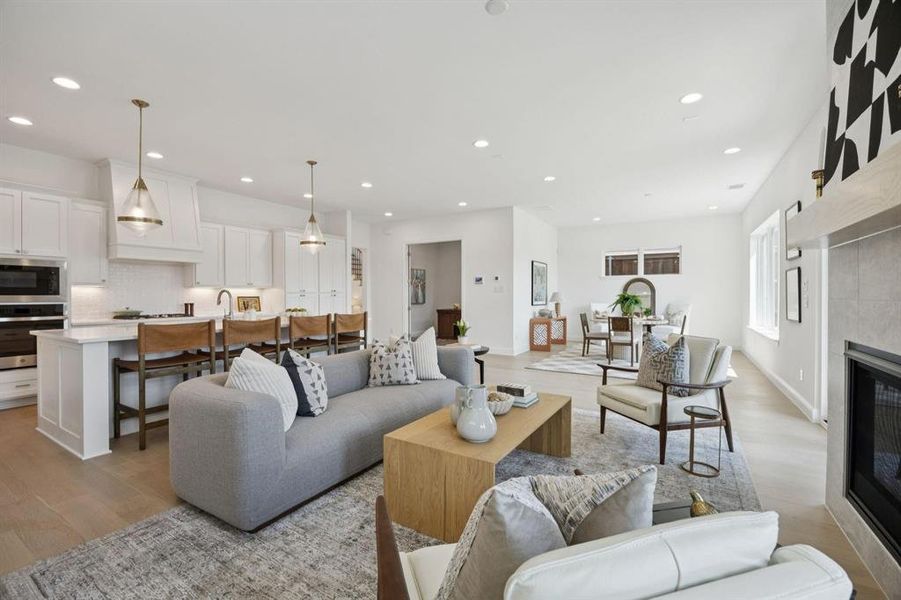 Expansive open-concept living area featuring light wood-finish flooring throughout, a modern fireplace with a light-toned tile surround, and a kitchen island with seating