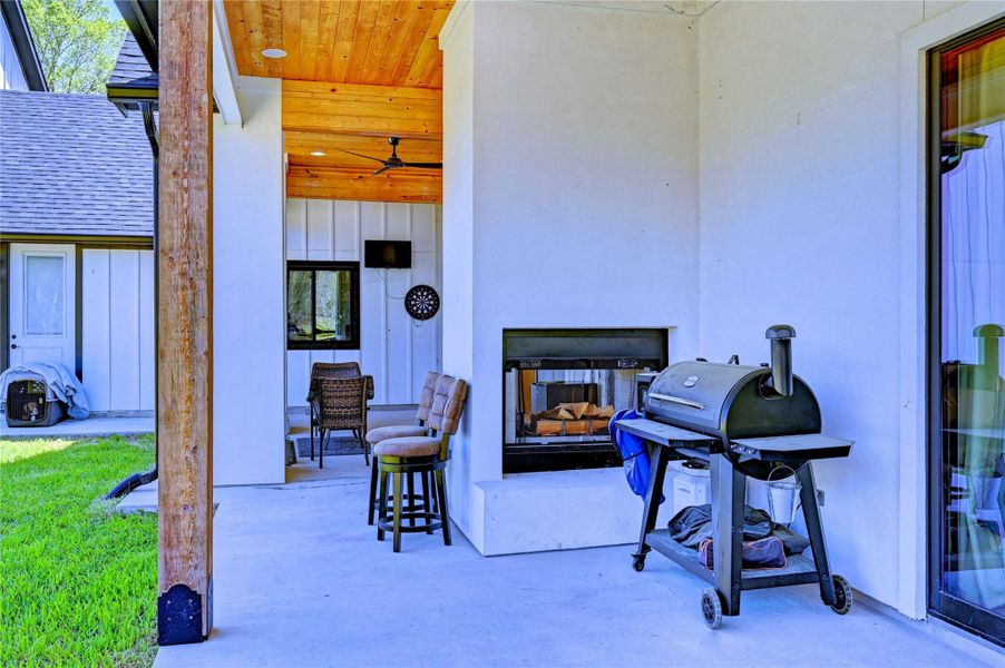 This photo showcases a covered outdoor patio with a cozy seating area and a built-in fireplace. It features a barbecue grill and is adjacent to a grassy yard, perfect for entertaining. The space has a warm wooden ceiling with a fan, enhancing comfort and style.