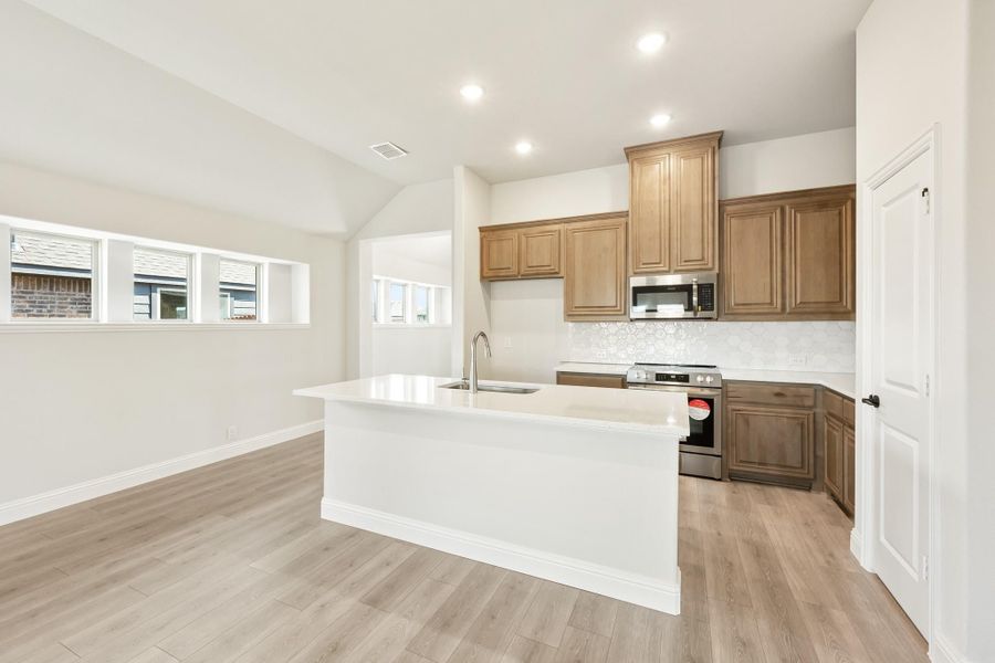 Kitchen with white island and sink, brown wood cabinets, stainless steel appliances, and light hardwood floors Kitchen with white island and sink, brown wood cabinets, stainless steel appliances, and light hardwood floors