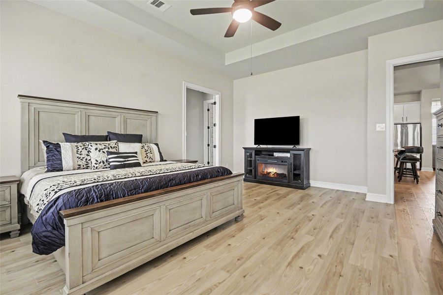 Spacious bedroom with light wood flooring, featuring a large bed and a cozy electric fireplace under a mounted TV. The room has neutral walls, a ceiling fan, and opens to a kitchen area. Spacious bedroom with light wood flooring, featuring a large bed and a cozy electric fireplace under a mounted TV. The room has neutral walls, a ceiling fan, and opens to a kitchen area.