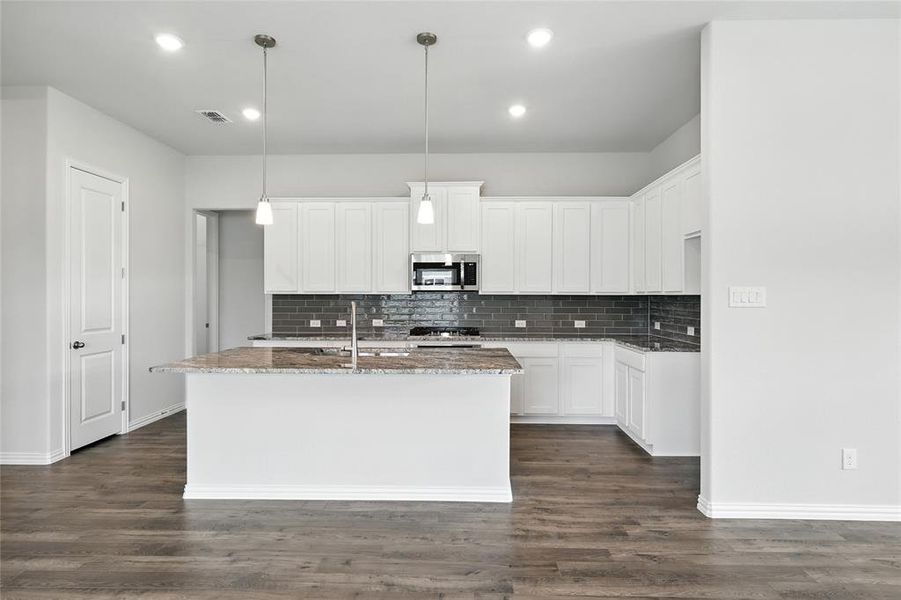 Kitchen featuring stainless steel microwave, stone counters, backsplash, a center island with sink, and dark wood-style flooring