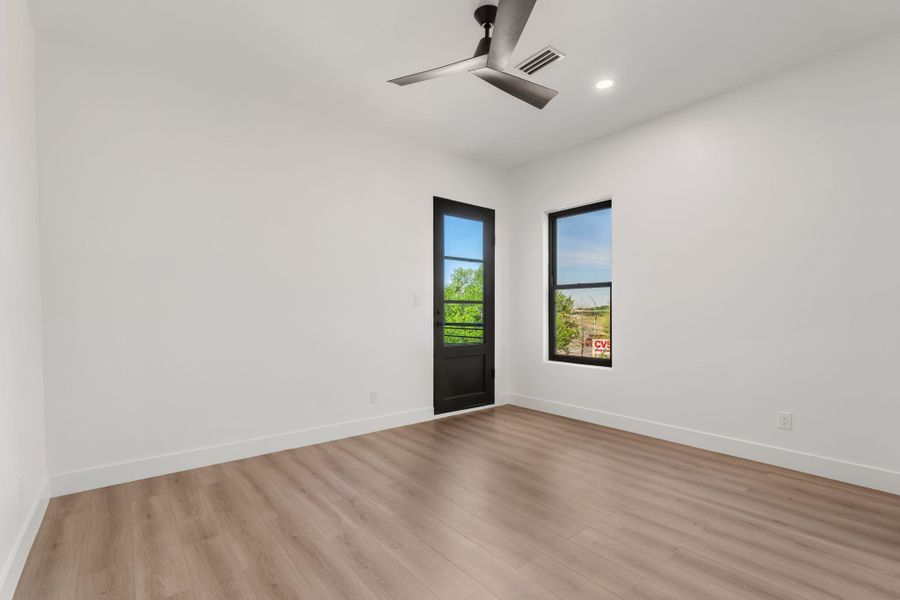 This secondary bedroom features a black-framed glass exterior door leading to the outdoor terrace and adjacent window that bring natural light in from two directions. Light vinyl plank flooring, a ceiling fan, and clean white walls carry the home's contemporary finish language throughout.