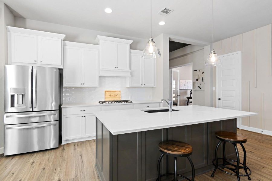 This modern kitchen features a sleek design with white cabinetry, stainless steel appliances, and a central island with a sink. The island offers seating with two stools and is illuminated by stylish pendant lights. The space has a clean, bright feel with light wood flooring.